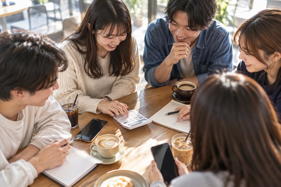 カフェのテーブルで電卓とスマホを使い、卒業旅行の費用を割り勘で計算する大学生グループ
